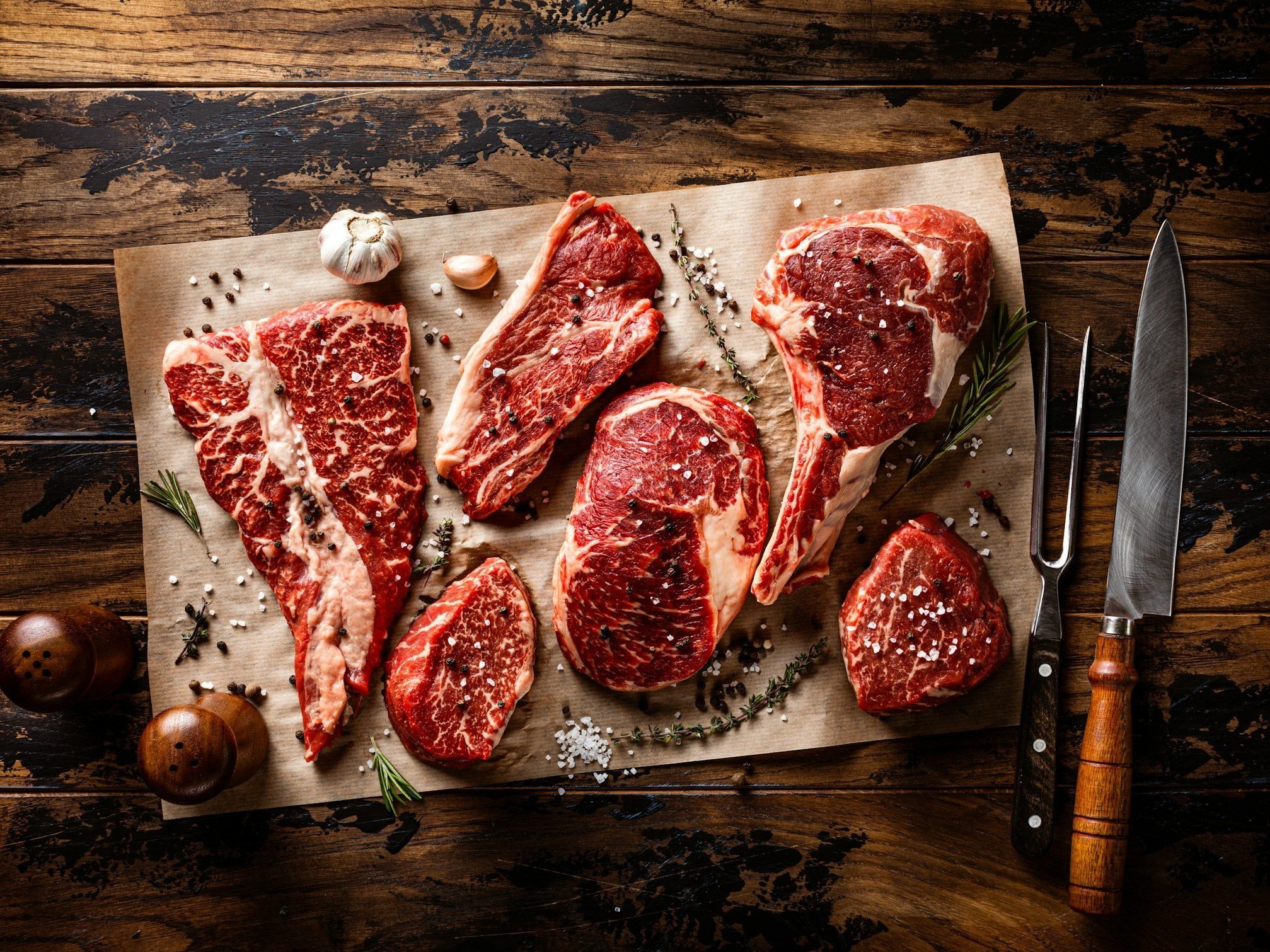 unsplash-image-JHWfH8V2INk (3) Assorted cuts of raw marbled beef steaks displayed on parchment paper, surrounded by garlic, herbs, and spices, with a knife and fork nearby on a rustic wooden surface.