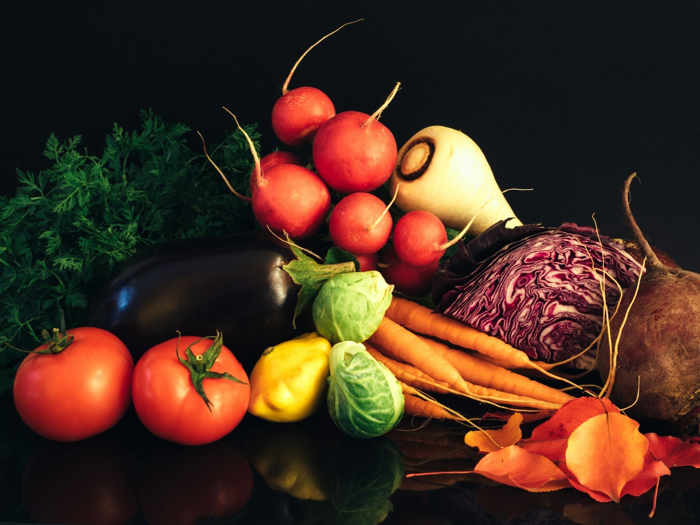 unsplash-image-KUZnfk-2DSQ Colorful assortment of fresh vegetables including tomatoes, radishes, carrots, eggplant, Brussels sprouts, yellow squash, purple cabbage, and beets, arranged artistically against a dark background, showcasing their vibrant colors and textures.